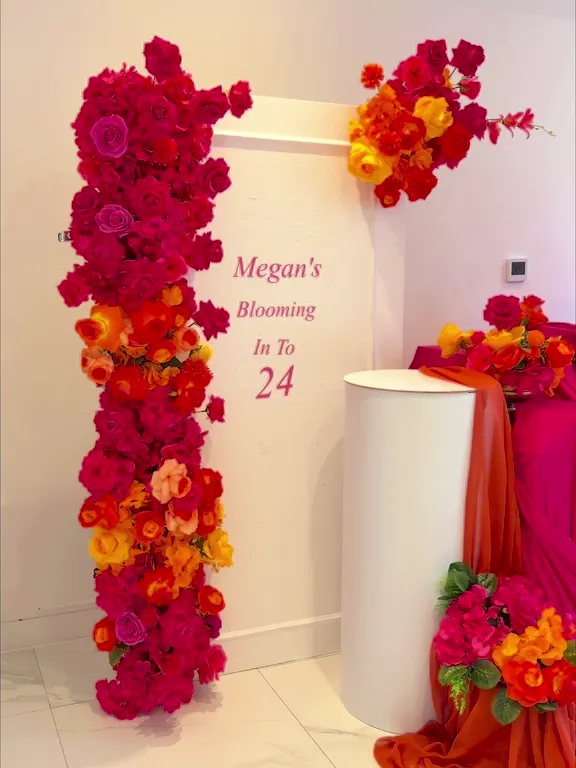 Birthday backdrop with a column of red, orange and yellow roses, a pink draped table and the words Blooming in to 24