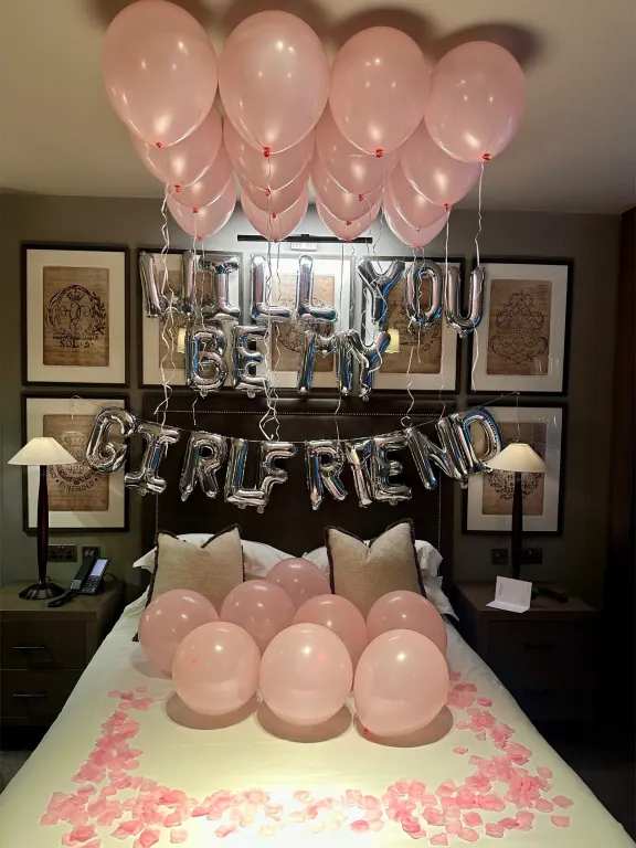 Hotel bed under pink balloons with Will you be my girlfriend in foil letters