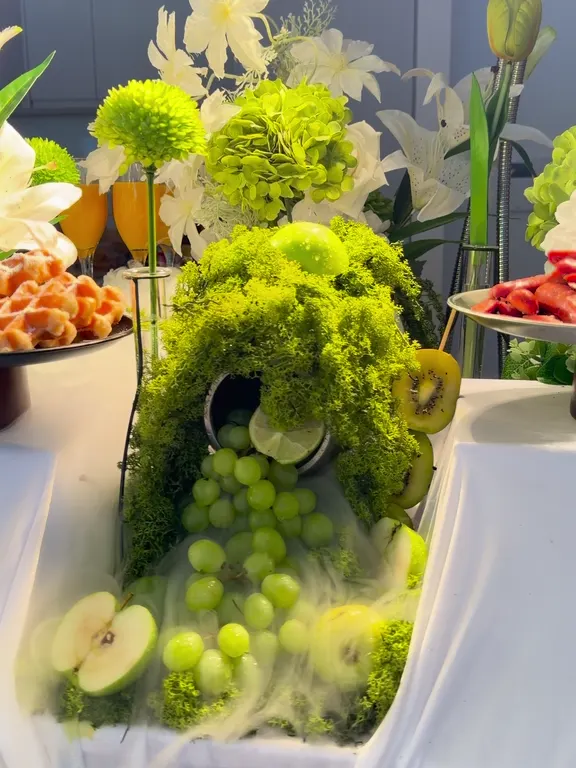 Grazing table with green grapes spilling from a mound of moss in drifting mist, under white lilies and green flowers