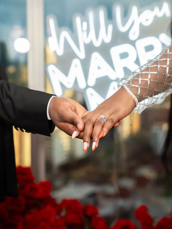 Two hands showing an engagement ring in front of a neon Will you marry me sign and red roses