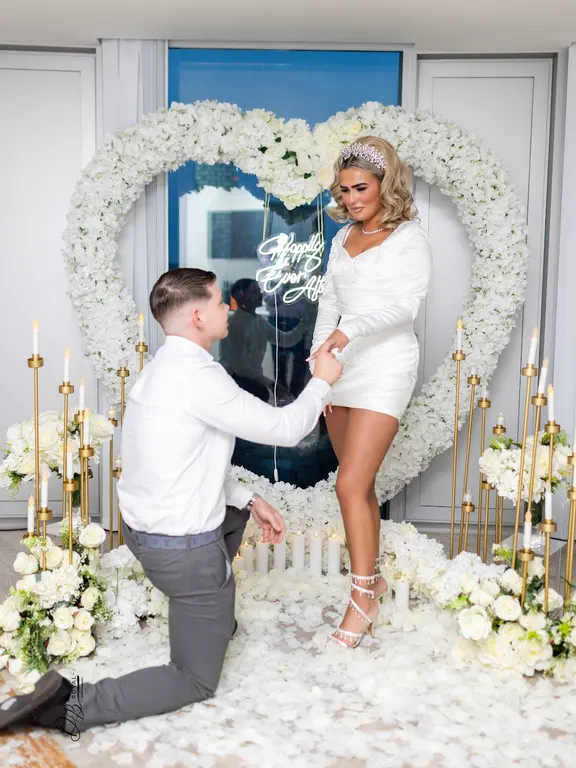 A man on one knee proposing in front of a tall heart of white flowers, surrounded by gold candle stands and white petals