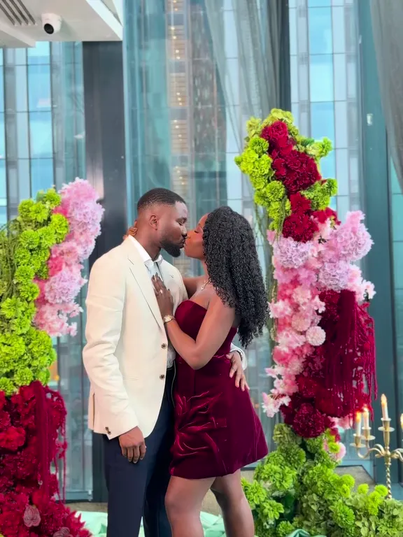 A couple kissing between tall flower pillars in a glass-walled room above the Manchester skyline